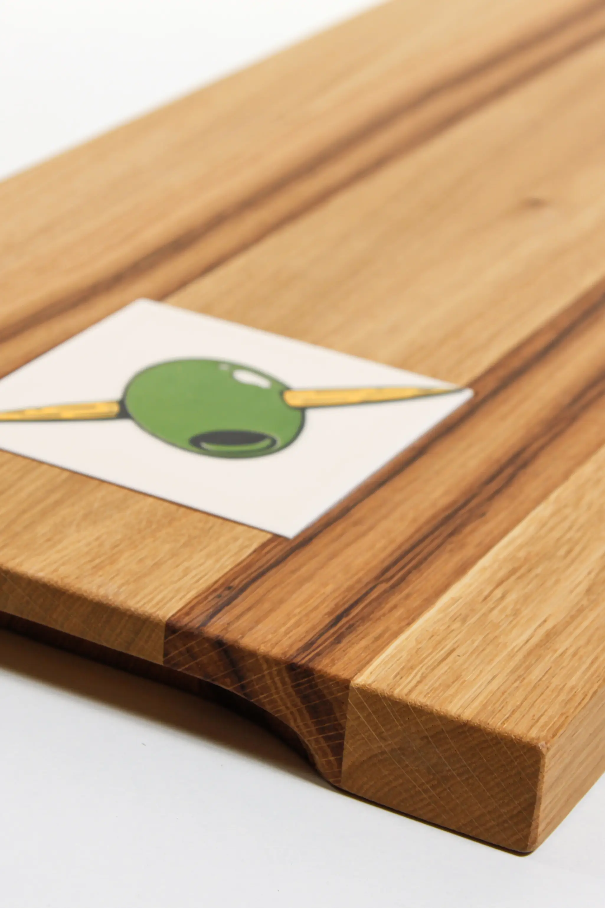Nahaufnahme eines Holzbretts mit einer eingelegten Kachel, die eine grüne Olive mit einem Zahnstocher zeigt.